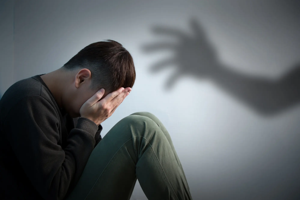 A distressed individual sits in a dimly lit space, haunted by the shadow of a hand, symbolizing PTSD treatment in Thousand Oaks, CA.