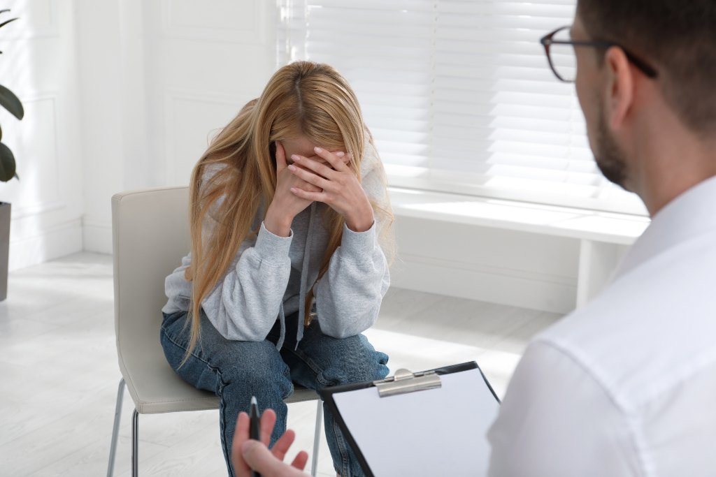 A woman sits in a chair, visibly distressed, with her head in her hands, while a therapist speaks with her. This scene highlights the importance of dual diagnosis treatment in Thousand Oaks, CA, where individuals receive care for both mental health and substance abuse issues.